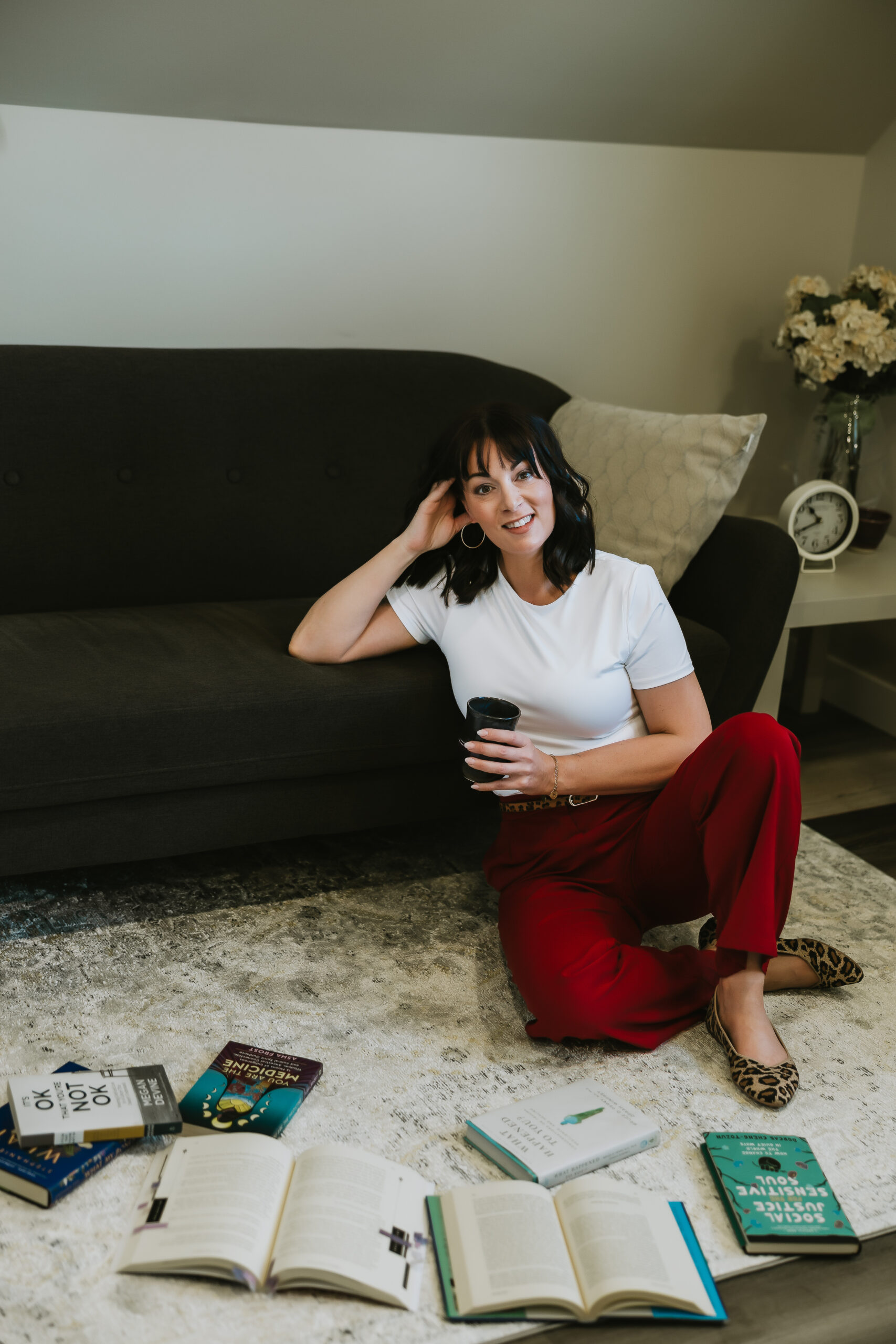 Stacey Stelle sitting on the floor in front of a couch with a welcoming smile, holding a coffee mug with books on the floor in front of her. 